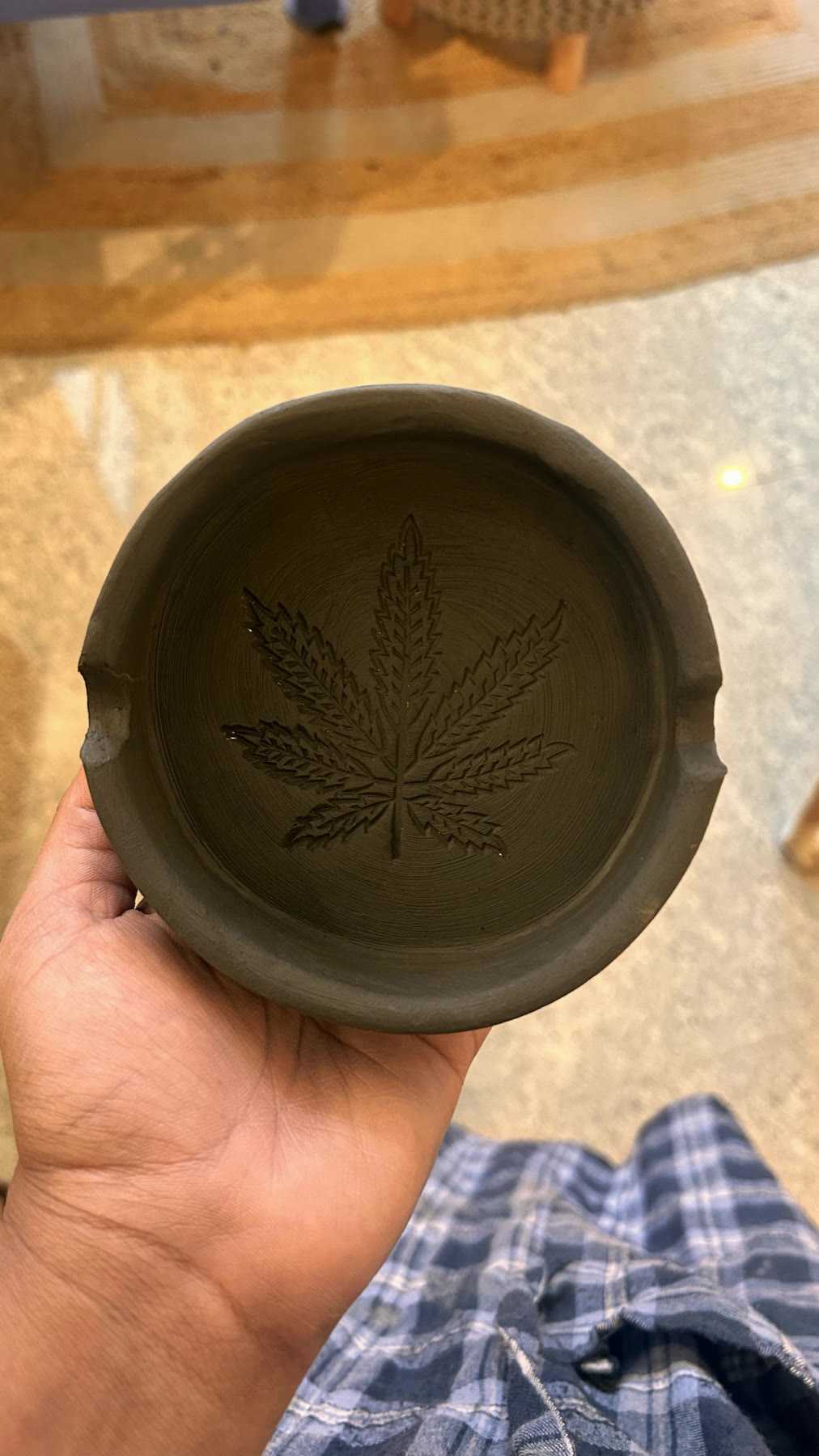 A hand holding a finished dark dish with a wild hemp leaf pressed into its base