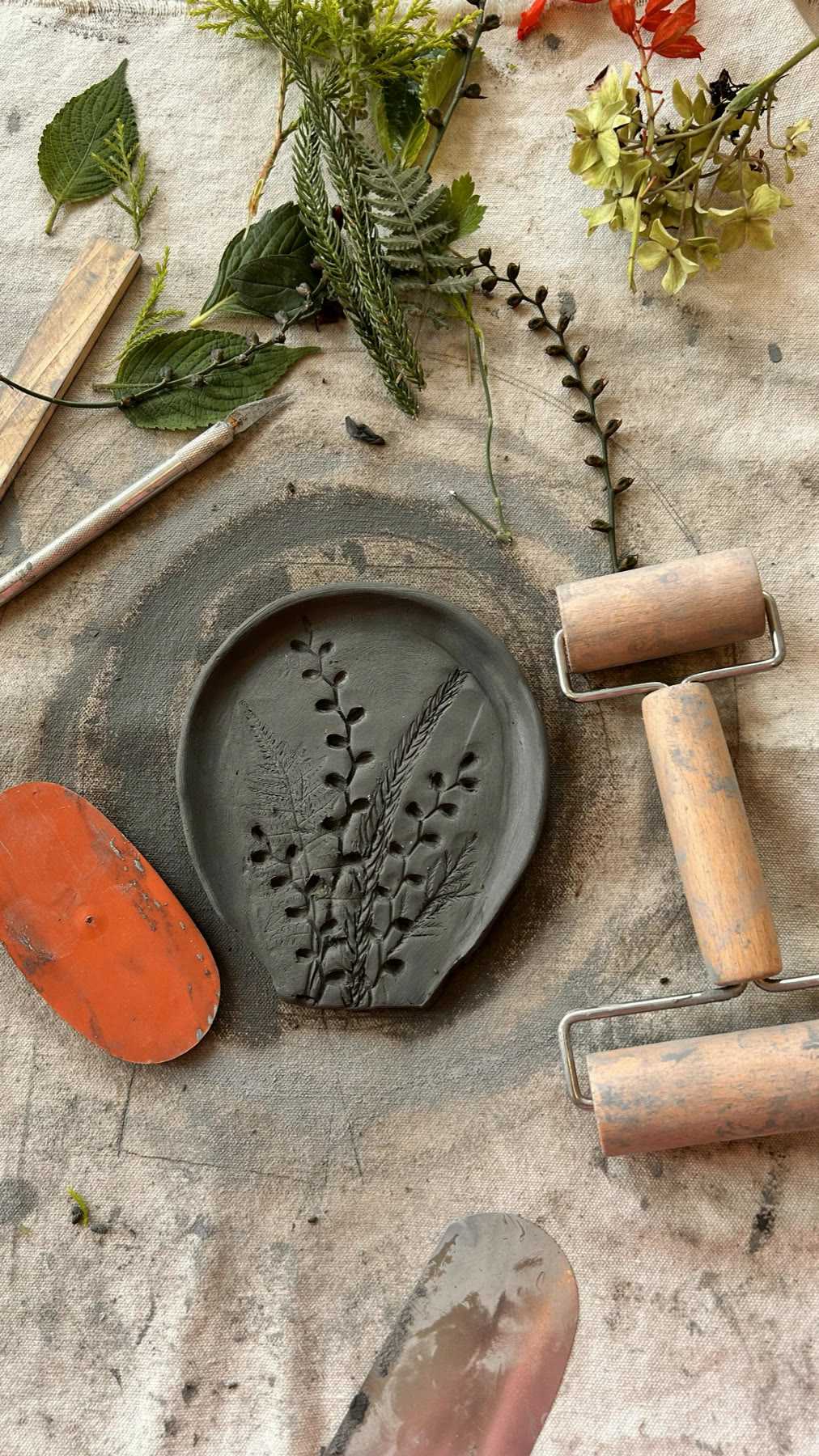 Foraged leaves and flowers laid around a clay slab printed with fern impressions