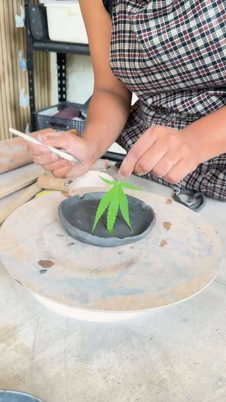 Hands pressing a fresh leaf into a dark clay dish with a wooden tool