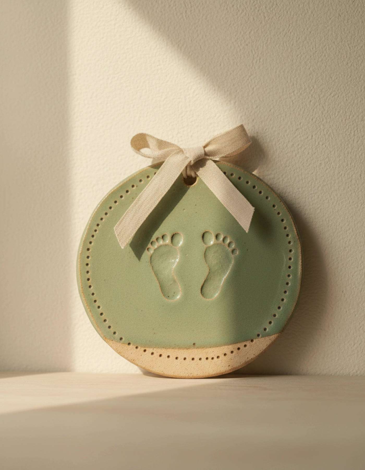 Illustration of a sage-green clay plaque with two baby footprints, hung from a ribbon