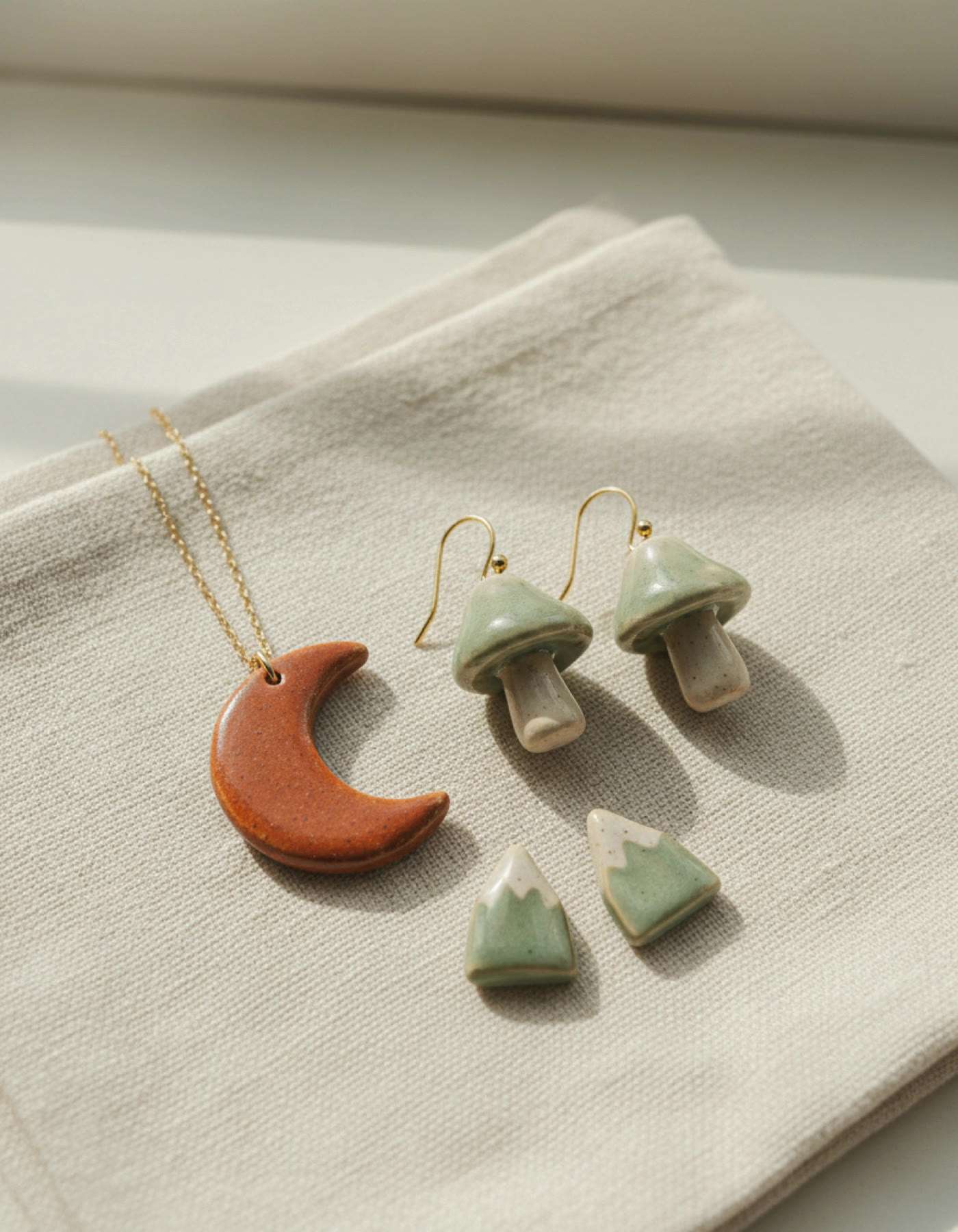 Illustration of handmade ceramic jewellery on linen: mushroom earrings, a moon pendant and mountain charms