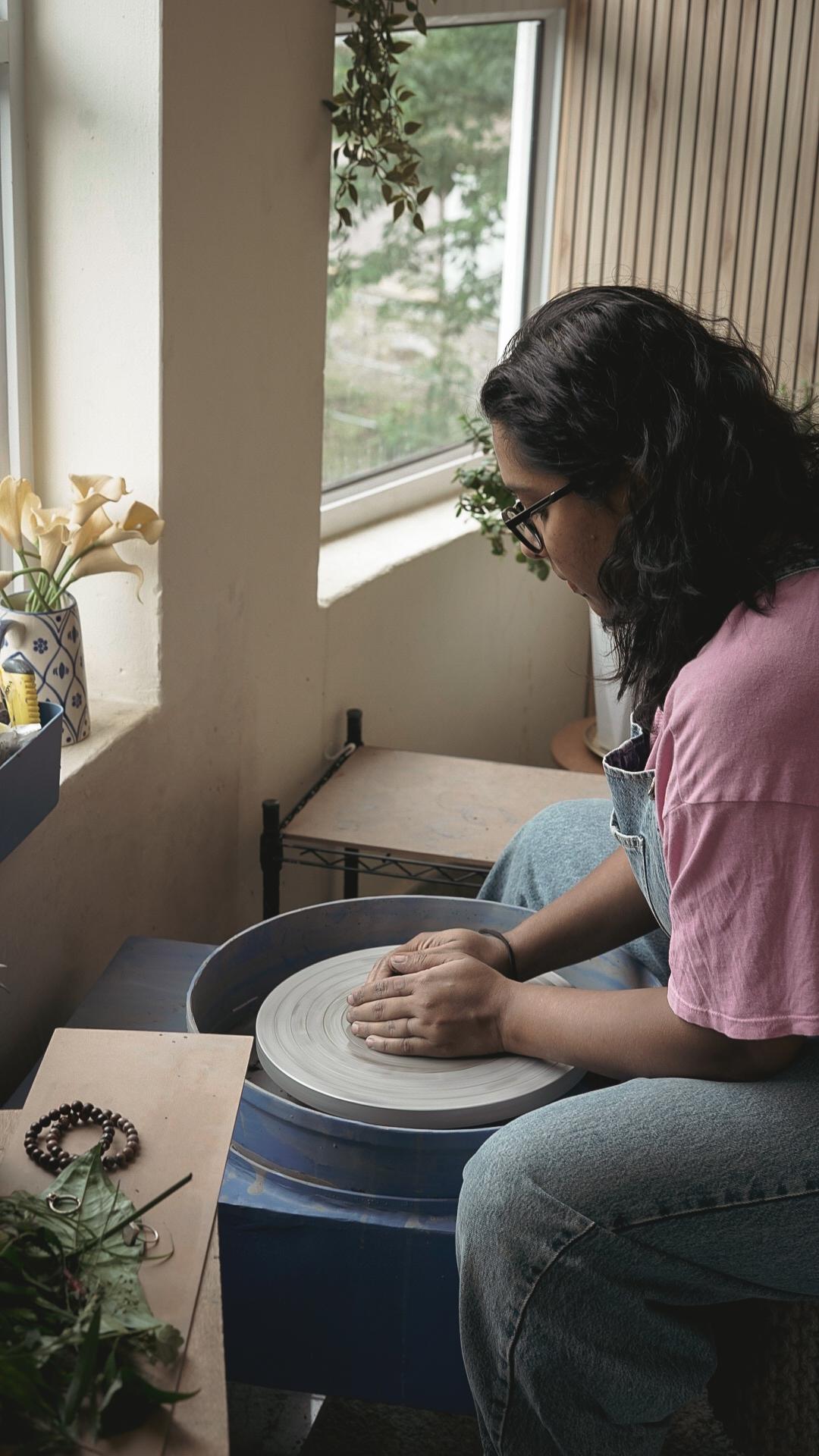 A potter at the wheel in the bright studio, greenery visible through the windows