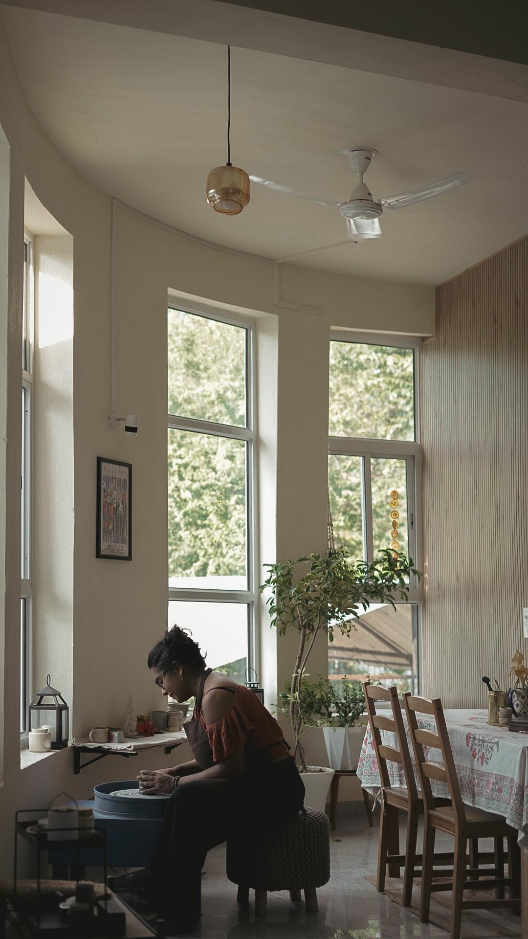 The studio seen from the doorway — pendant light, plants along the windows, Madhumita at the wheel in the corner