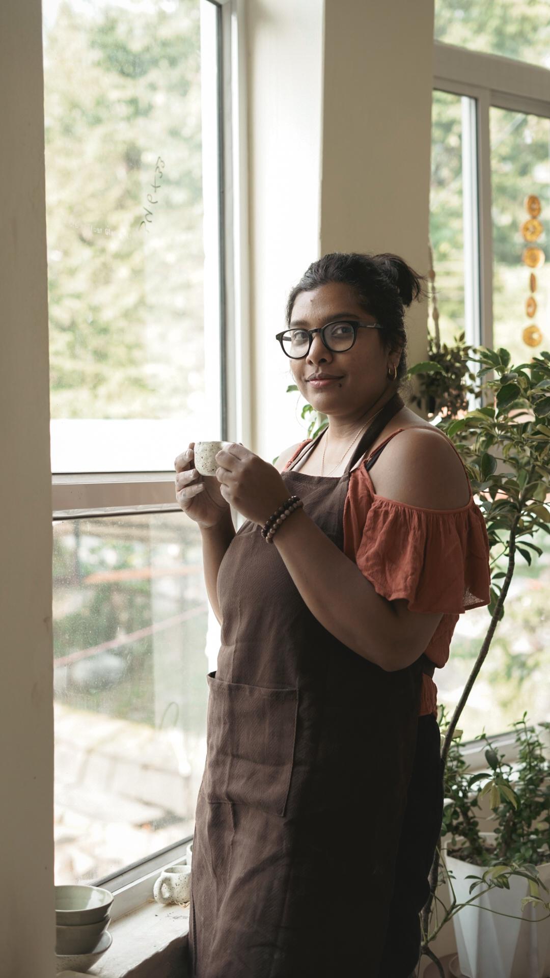 Madhumita Chatterjee holding a handmade cup in the sunlit studio, looking at the camera