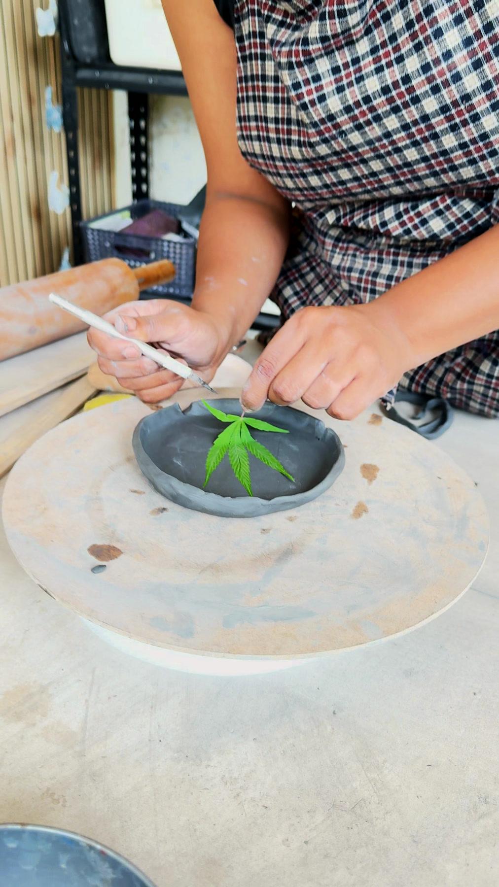 Hands pressing a fresh leaf into a dark clay dish, the studio window behind