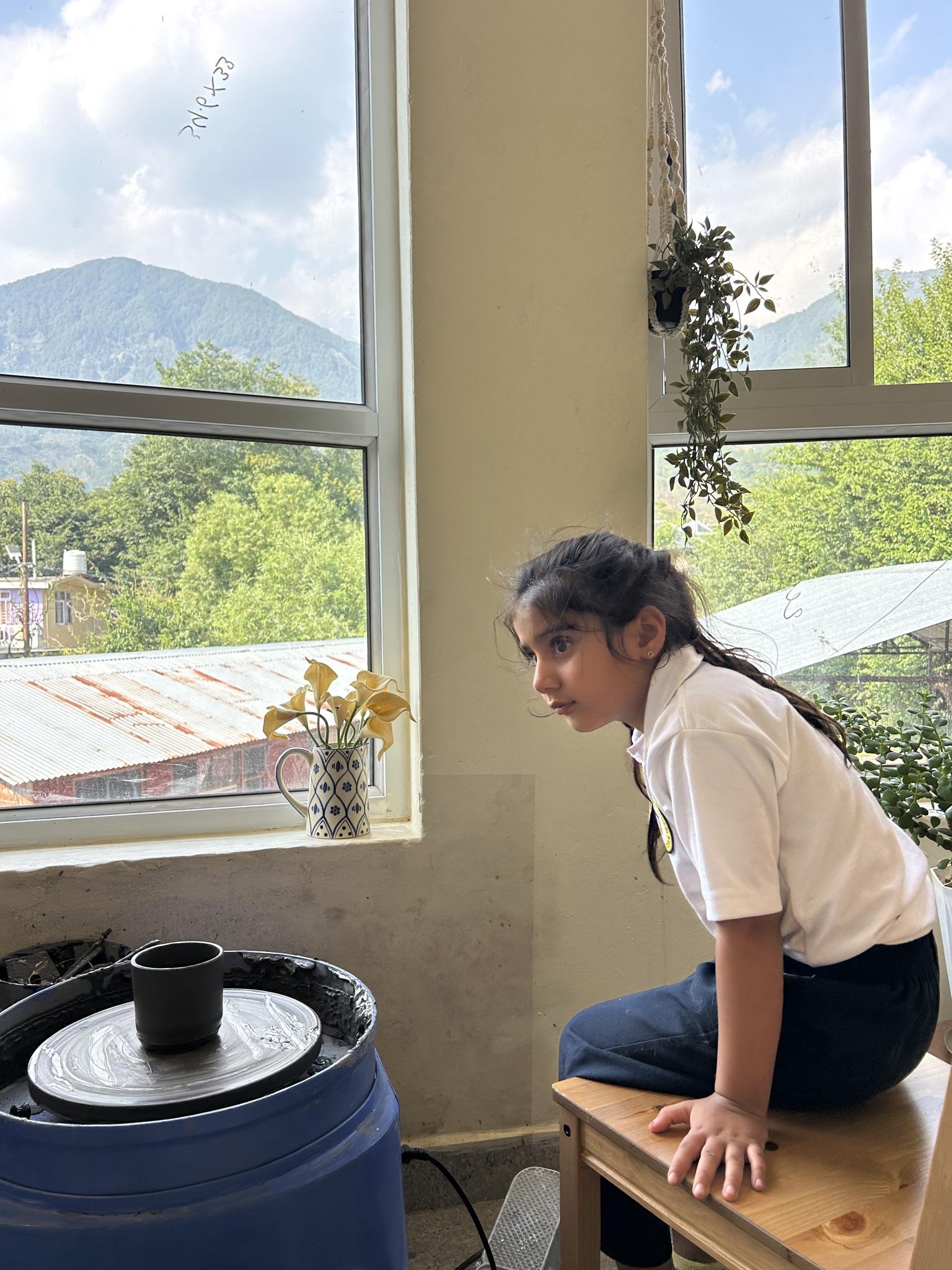 A student leaning in to watch a small dark cup turning on the wheel, the mountain and tin roofs of the village through the window behind her