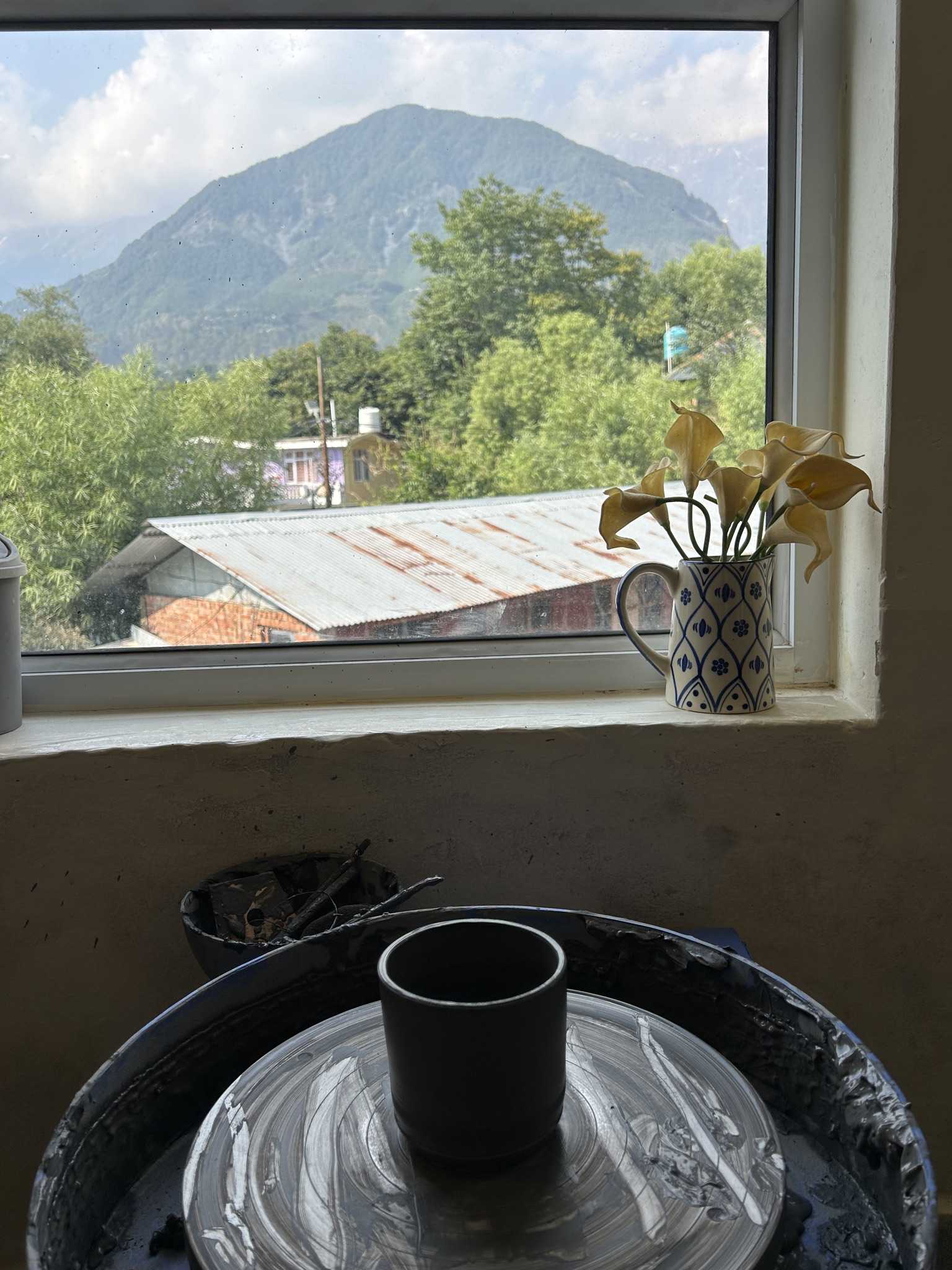 A dark cup standing on a pottery wheel, the mountain and tin roofs framed in the window behind