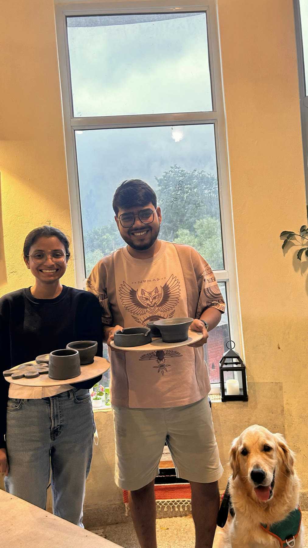 Two guests holding a board of the cups and bowls they made, the studio dog sitting beside them