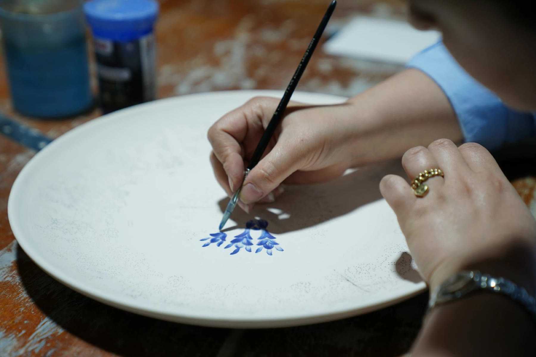 A hand painting a small blue floral motif onto a white ceramic plate