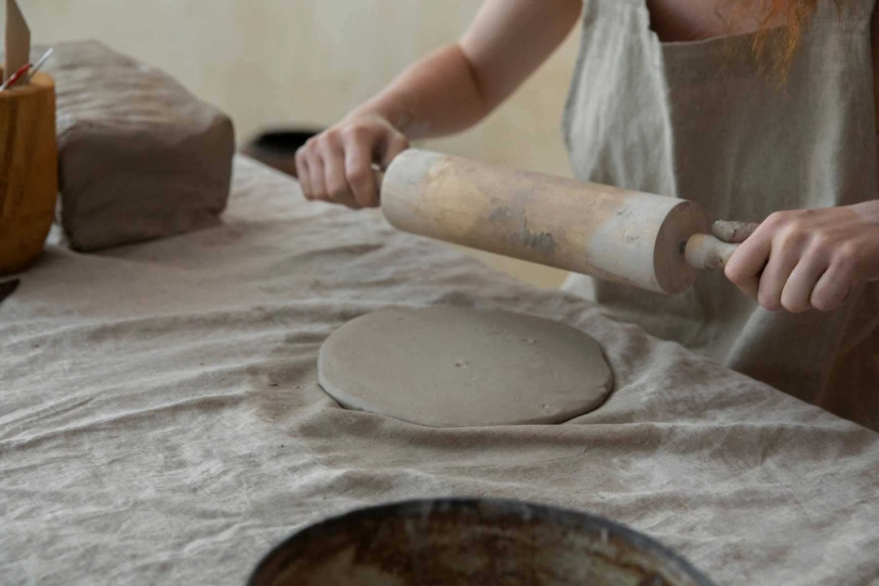 A rolling pin flattening a round clay slab on a canvas-covered table
