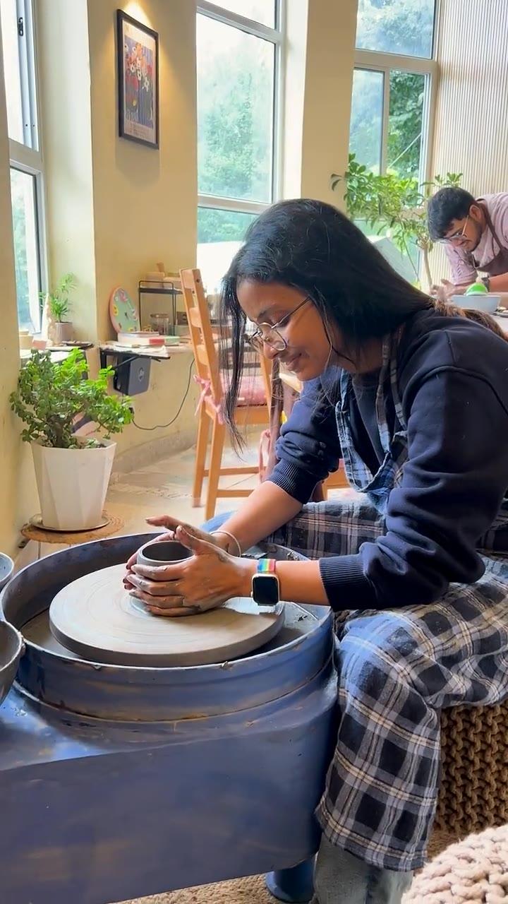 A guest at the pottery wheel, smiling, with the valley visible through the studio windows behind her