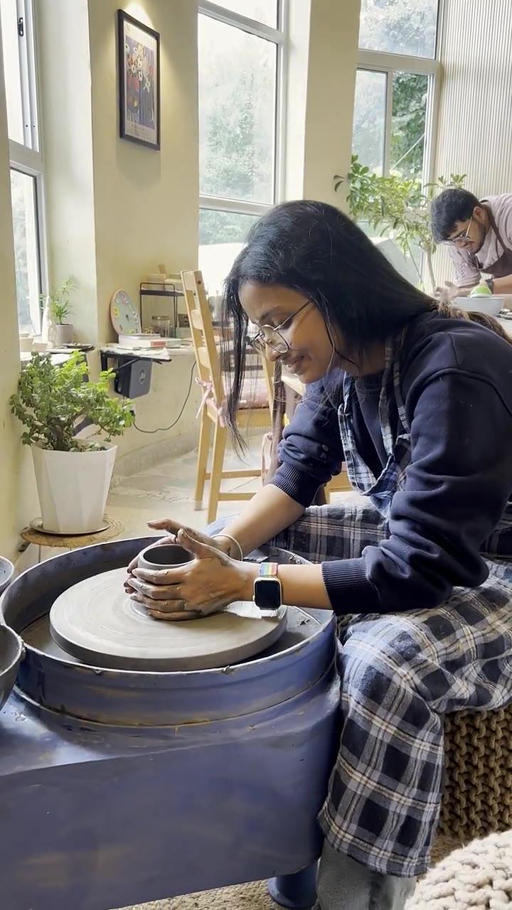 A guest at the pottery wheel, smiling, with the valley visible through the studio windows behind her