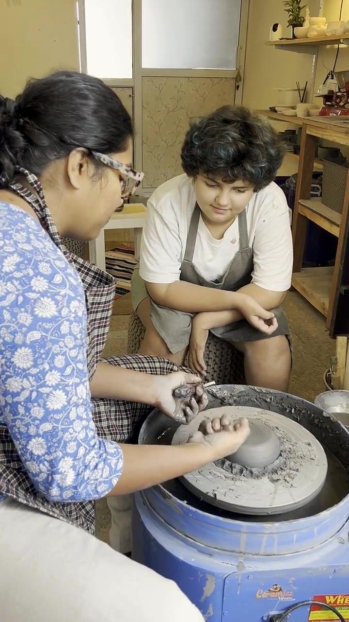 Madhumita with her hands over a young student's hands, steadying a pot on the wheel